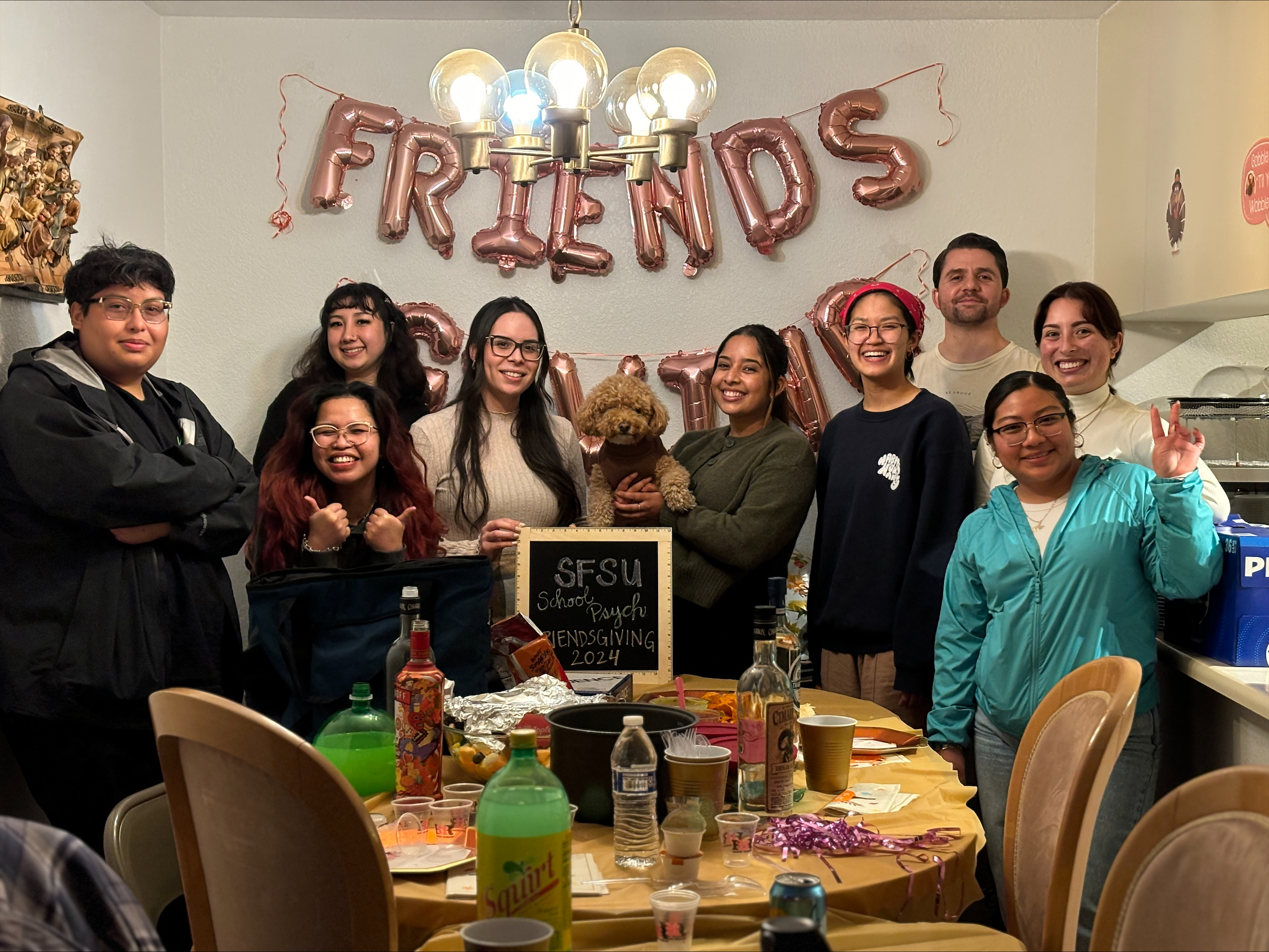 School Psychology Fall 2022 Cohort Friendsgiving. People gathered around a table with balloons that spell out "Friends" on the wall. Someone in the middle is holding a dog.